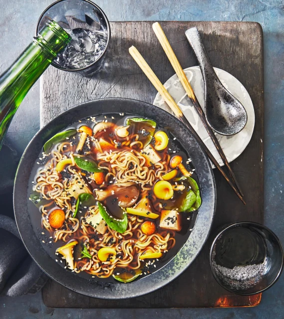 Overhead shot of noodle soup with vegetables and mushrooms being poured from a green bottle, accompanied by chopsticks and a spoon.