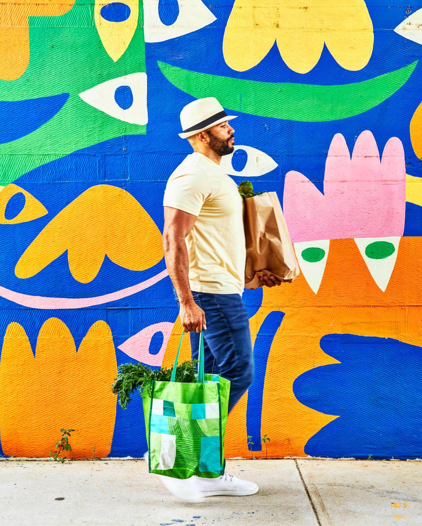 Man with paper bag and tote walks past colorful geometric mural in jeans, t-shirt, and hat on sidewalk in urban environment.