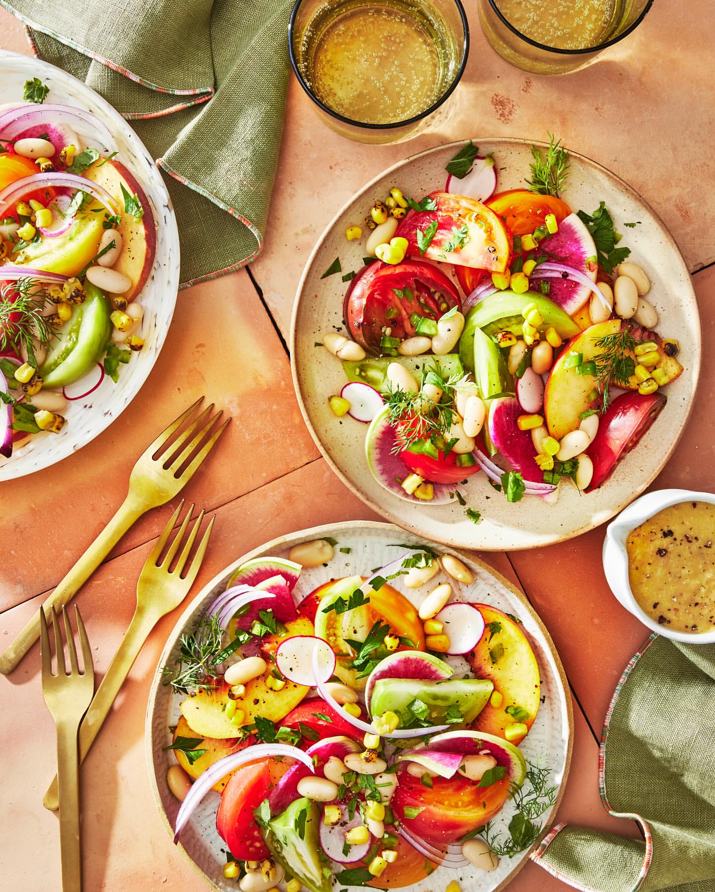 Overhead shot of colorful tomato salads with white beans, corn, and red onion on plates with forks, featuring glasses of beverage.