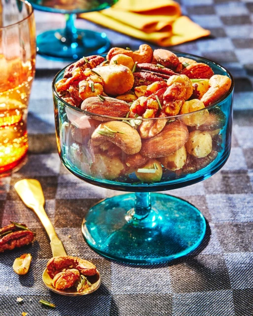 Assortment of nuts served in turquoise glass pedestal bowl with golden spoon on grey checkered tablecloth, studio shot.