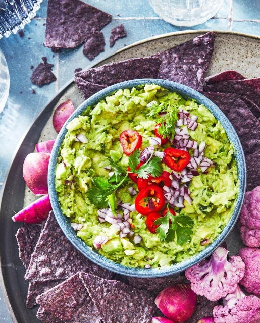 Overhead shot of homemade guacamole in a blue bowl, garnished with red chili, chopped onion, and cilantro, served with blue corn chips.