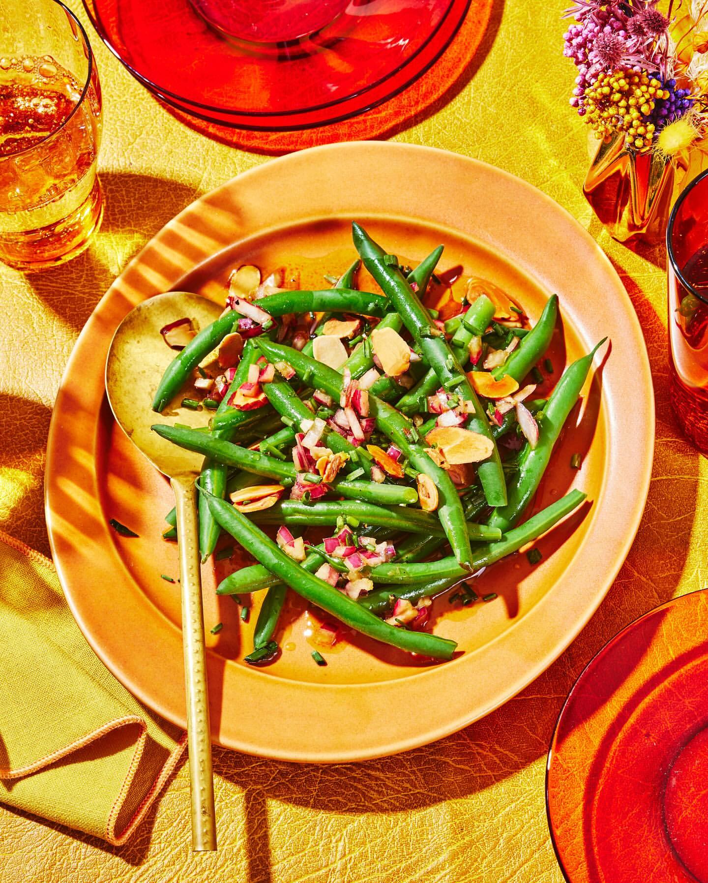 Overhead shot of a green bean salad on a orange plate with almonds and red onion on a yellow tablecloth next to drinks.
