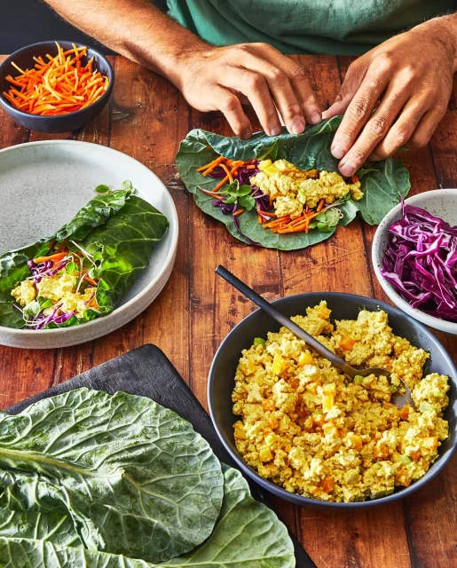 Collard green wraps being assembled on a wooden table with bowls of ingredients like shredded carrots, cabbage, and filling.