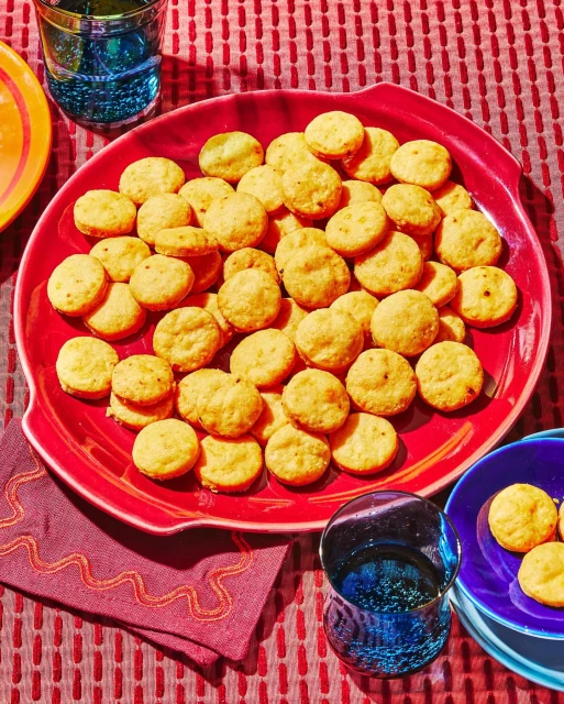 Overhead shot of a red plate filled with cheese crackers alongside a purple napkin, blue drinking glass, and red patterned tablecloth.