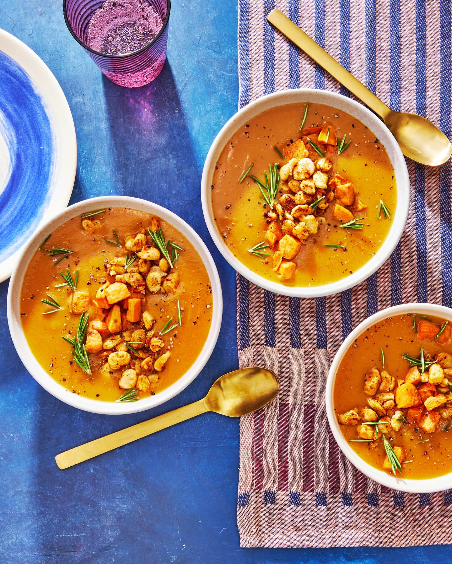 Overhead shot of three bowls of butternut squash soup with rosemary and croutons on a striped towel on a blue surface.
