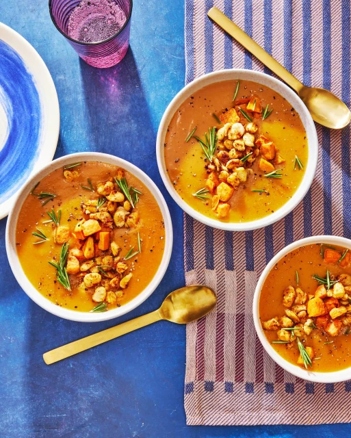 Overhead shot of three bowls of butternut squash soup with rosemary and croutons on a striped towel on a blue surface.