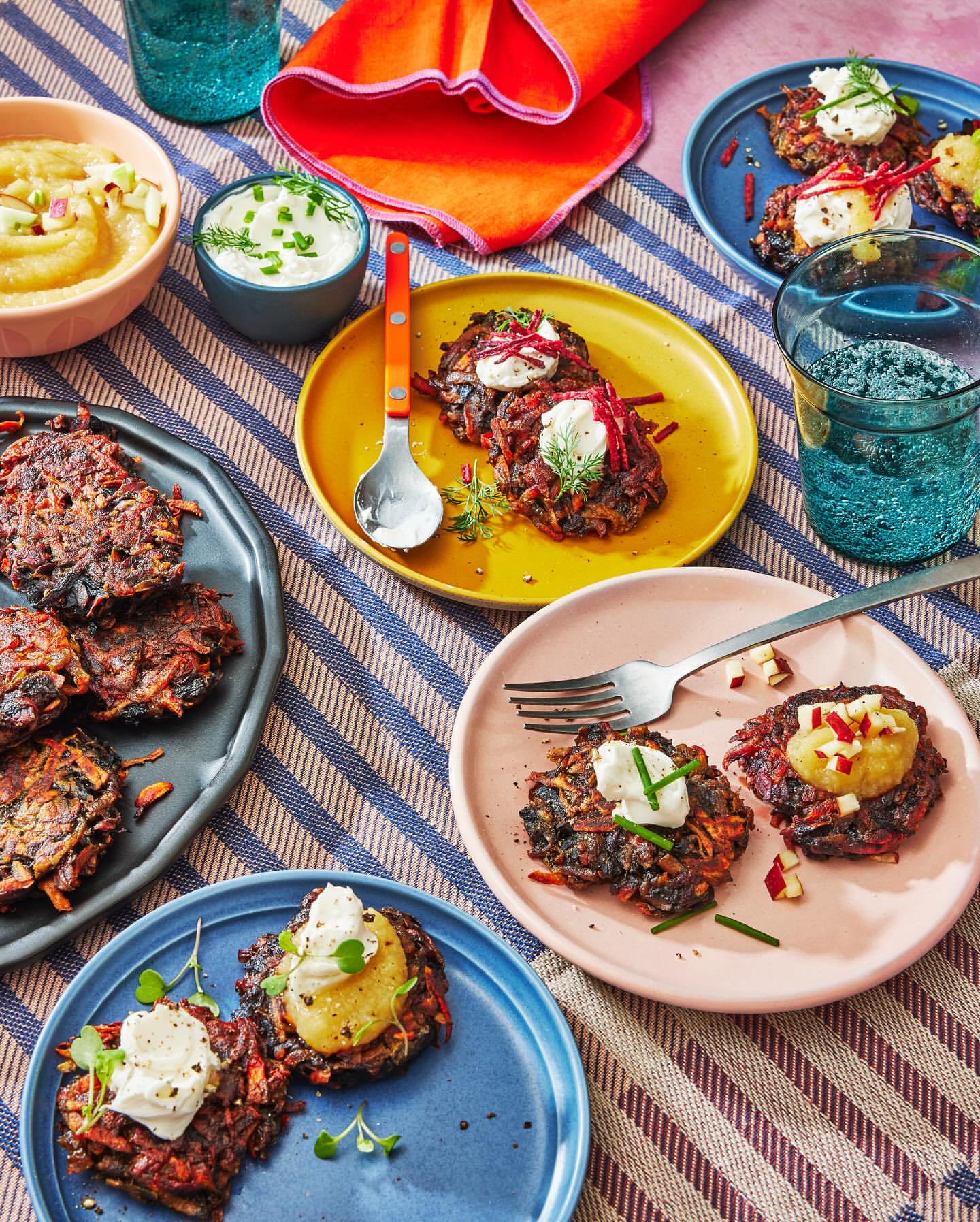 Overhead shot of colorful plates with vegetable fritters topped with cream and applesauce, arranged on a striped tablecloth.