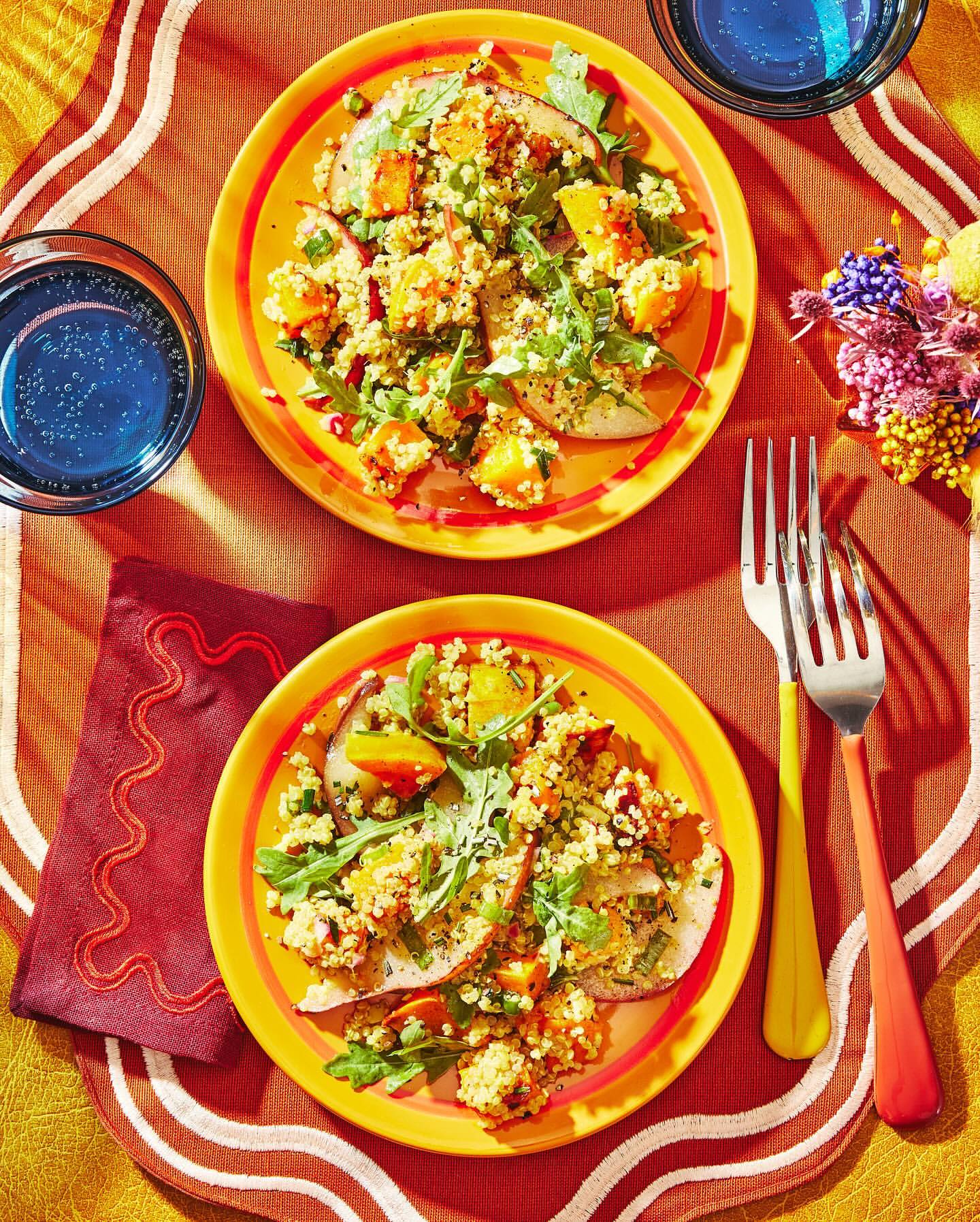 Overhead shot of two yellow plates with pear salads topped with grains, squash, and greens, served with blue drinks and silverware.