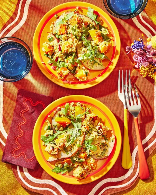 Overhead shot of two yellow plates with pear salads topped with grains, squash, and greens, served with blue drinks and silverware.