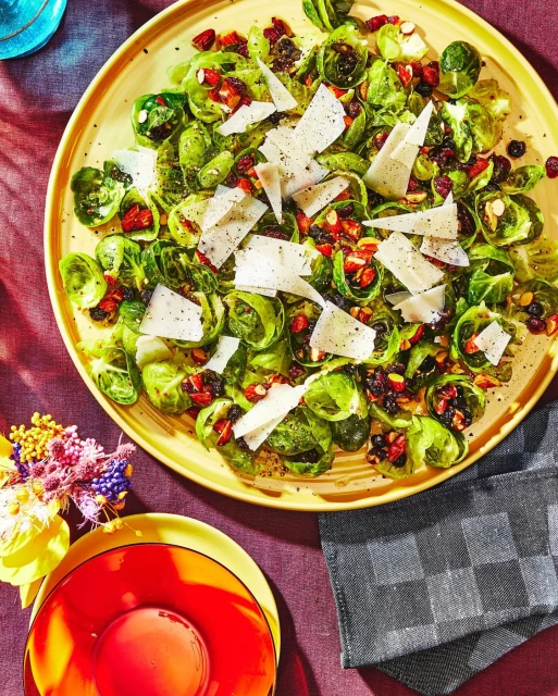 Overhead shot of a yellow plate with brussel sprouts salad, topped with cranberries and cheese, placed on a maroon table cloth.