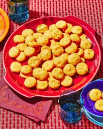 Overhead food photography of golden crackers piled on a large red serving platter on a patterned red textile background with blue glassware