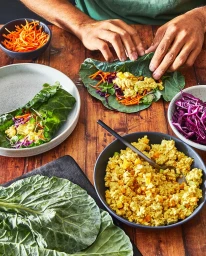Close-up of hands rolling a green leaf wrap filled with shredded carrots and yellow filling on a wooden table.