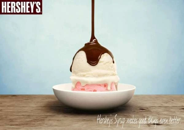 Chocolate syrup being poured over a stack of vanilla and strawberry ice cream in a white bowl on a wood surface against a light blue background.