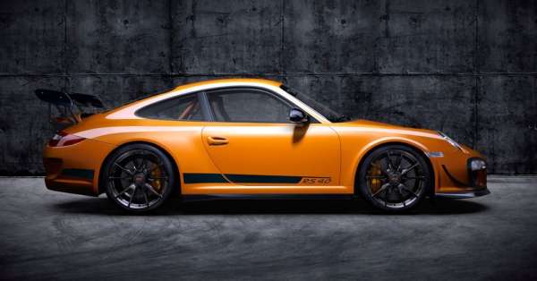 Orange Porsche 911 RS 4.0 with black wheels and spoiler parked indoors against a grey textured background.