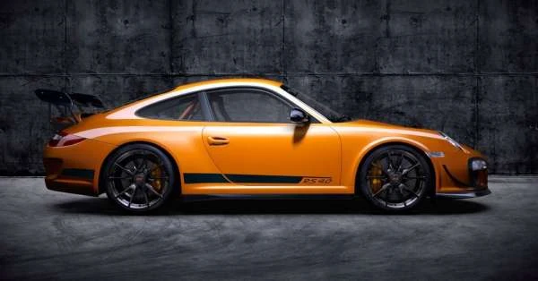 Orange Porsche 911 RS 4.0 with black wheels and spoiler parked indoors against a grey textured background.