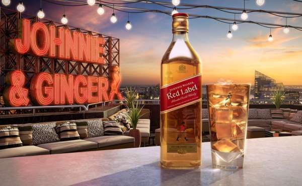 A bottle of Johnnie Walker Red Label whisky is featured with a glass of ice against a city skyline at sunset, under string lights.