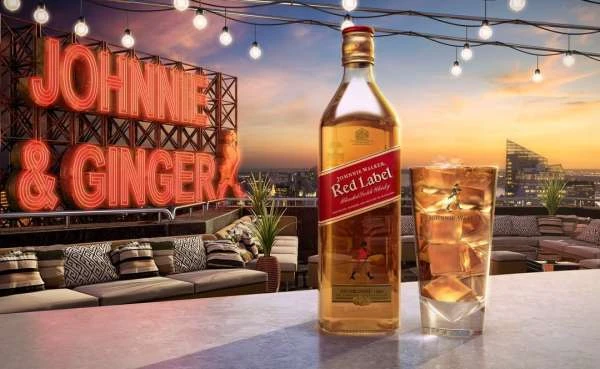 A bottle of Johnnie Walker Red Label whisky is featured with a glass of ice against a city skyline at sunset, under string lights.