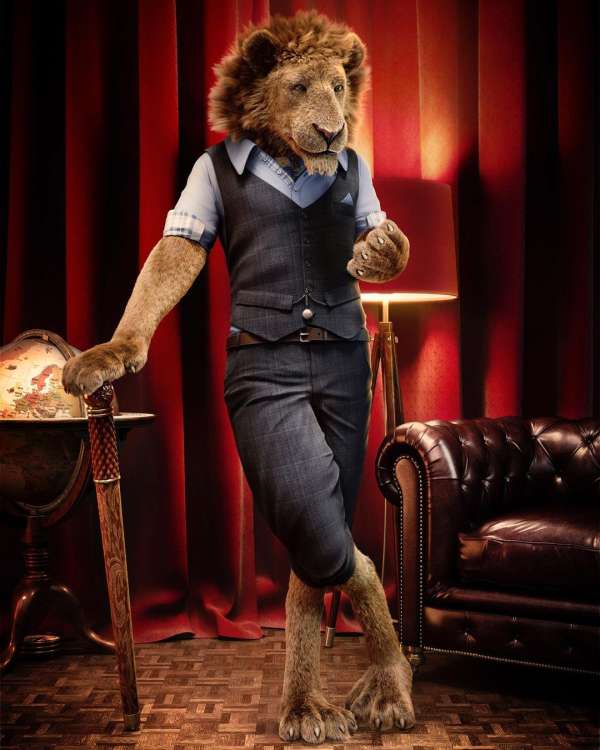 An anthropomorphic lion stands in formal wear leaning on a cane next to a globe and a chair against a curtained wall.