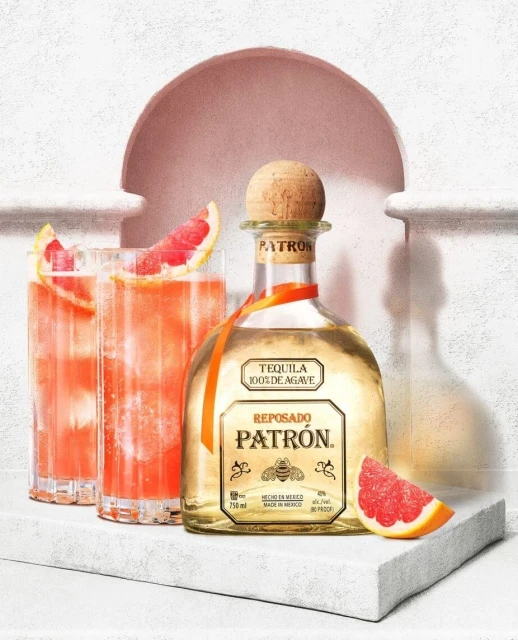 Patron Reposado tequila bottle with two cocktails garnished with grapefruit slices on a white display, against a plaster background.