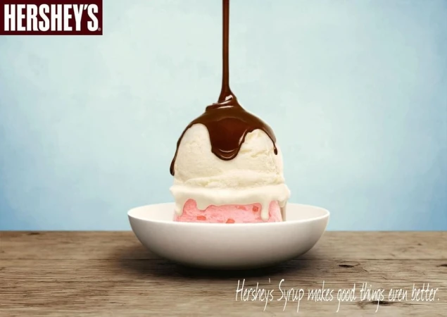 White and pink ice cream scoops are topped with Hershey's chocolate syrup in a white bowl on a wooden table against a blue background.