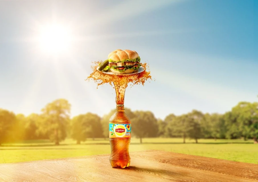 Stylized advertising shot of a Lipton Iced Tea bottle under a hamburger on a plate against a summer sky and park background.