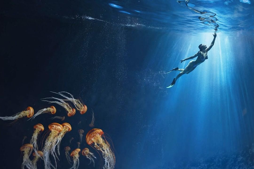 Woman swims underwater in ocean with jellyfish, sunlight filters from above, creating a scene of ethereal beauty and tranquility.
