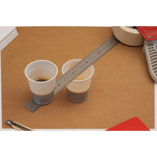 Two plastic cups sit on a brown surface beside a metal ruler, tape dispenser, utility knife, and red plastic smoother tool.