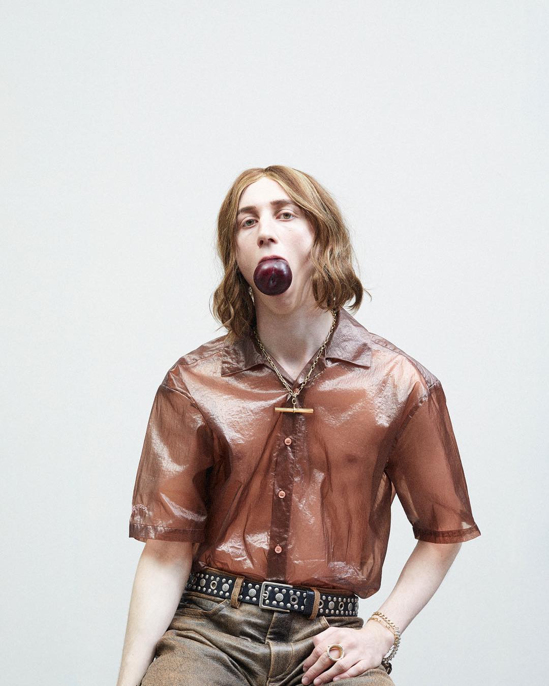 Stylish individual with brown hair poses in a semi-sheer bronze shirt, accessorized with jewelry and a black studded belt against a white backdrop.