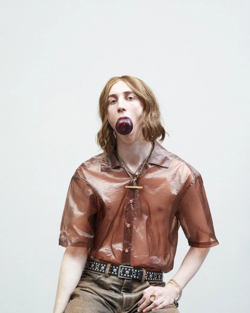 Stylish individual with brown hair poses in a semi-sheer bronze shirt, accessorized with jewelry and a black studded belt against a white backdrop.