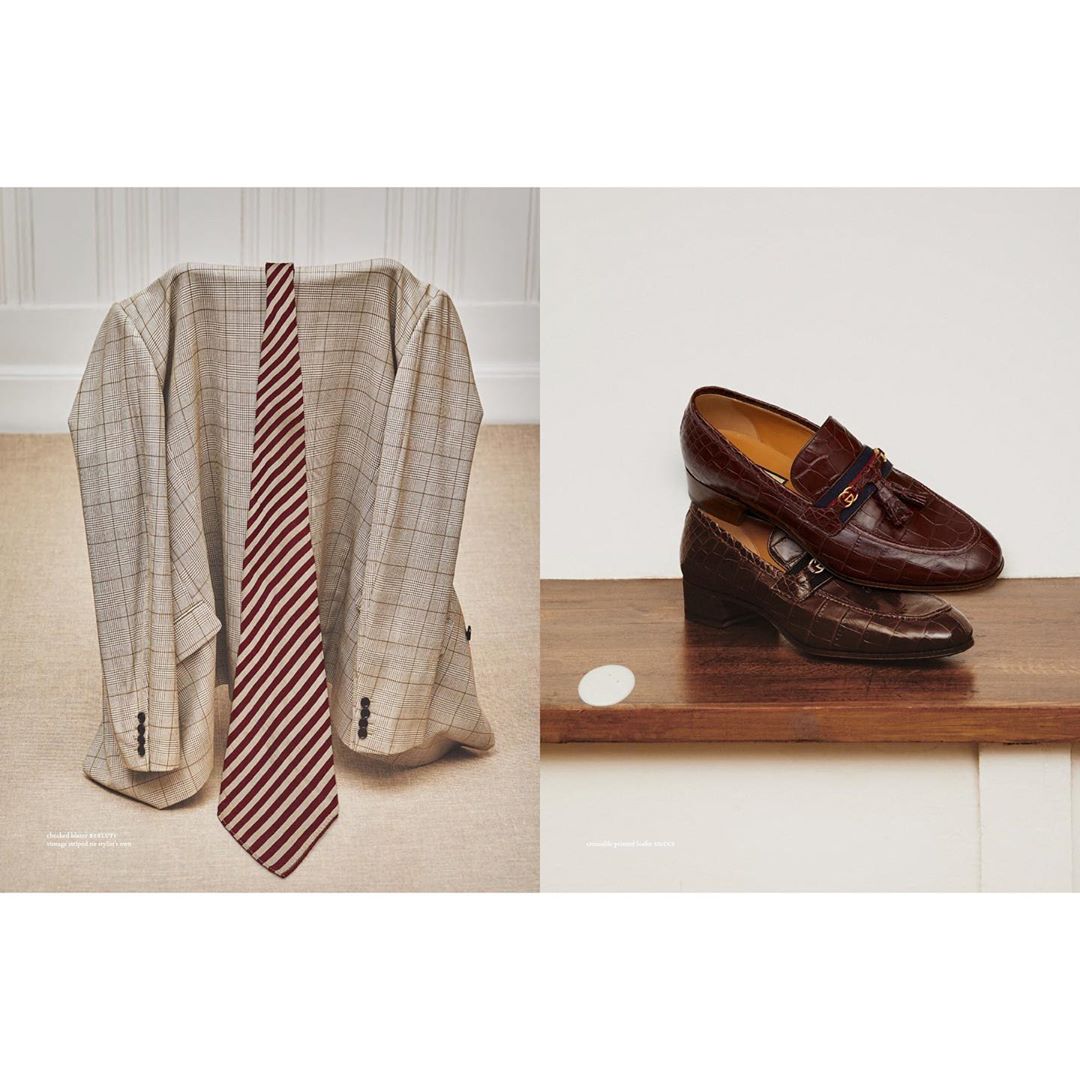 Beige plaid suit jacket and red striped tie next to a stack of brown leather loafers on a wooden shelf in front of a white wall.