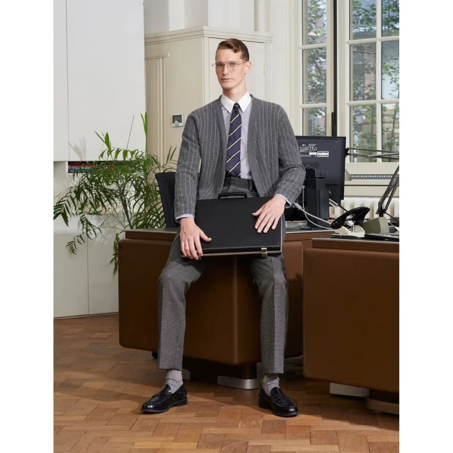Man in pinstripe suit sitting on a desk in an office. He wears glasses and holds a black briefcase in his hands.
