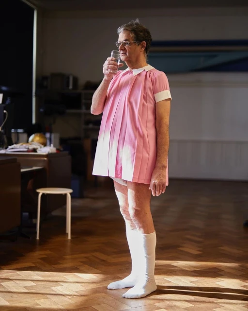 Older man in a short pink dress and long white socks is drinking water. The photo is taken inside a building on a wood floor.