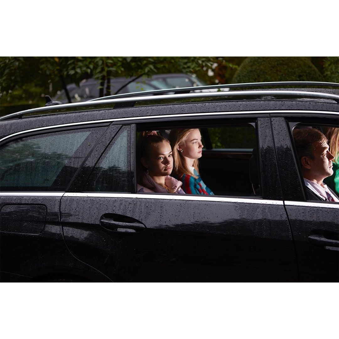 Two fair-skinned teenage girls and an adult male are seen in a dark SUV through the passenger window, under overcast daylight conditions.