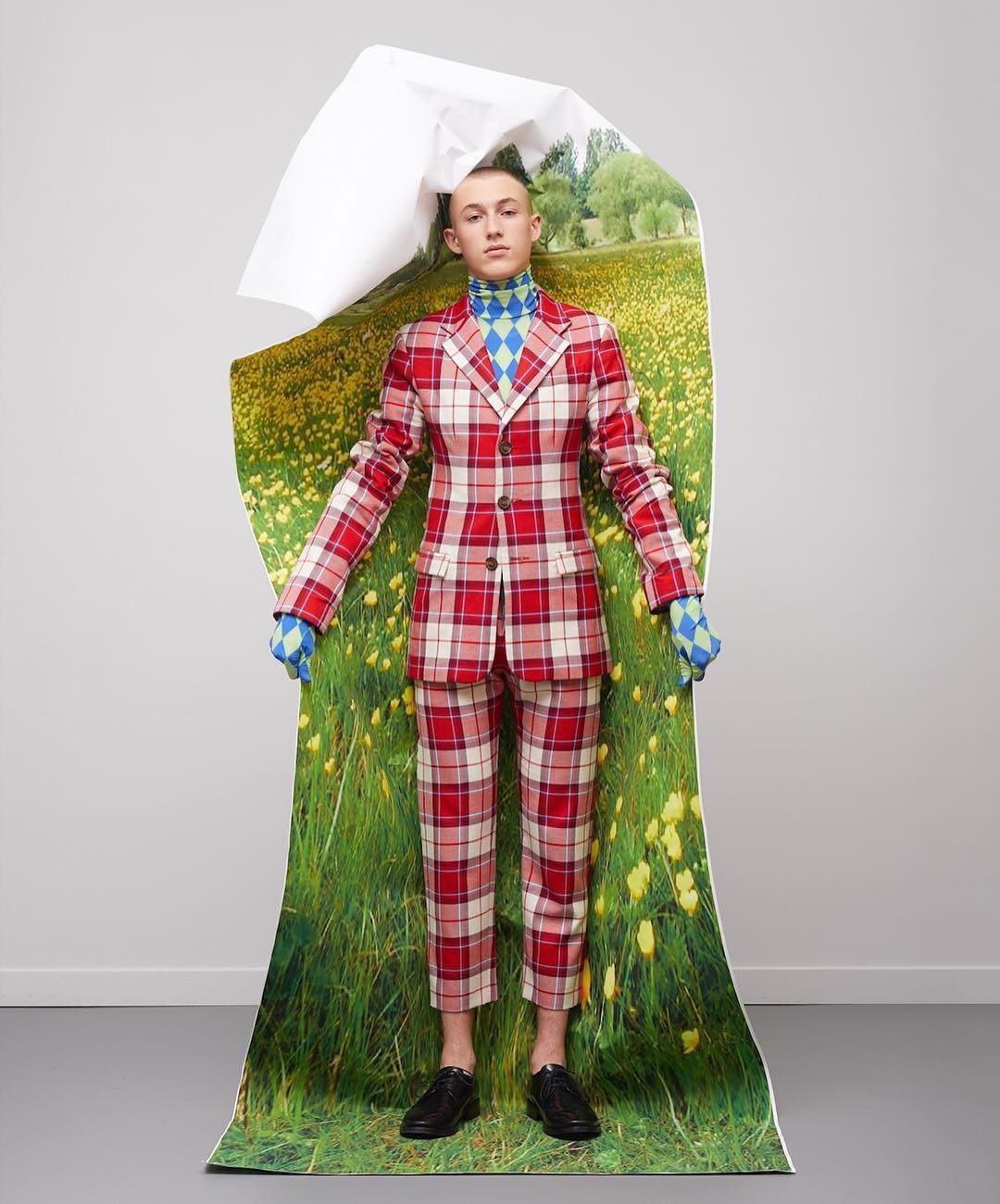 A person stands indoors wearing a red plaid suit, a blue argyle turtleneck, and black shoes. Behind them is a torn field image.