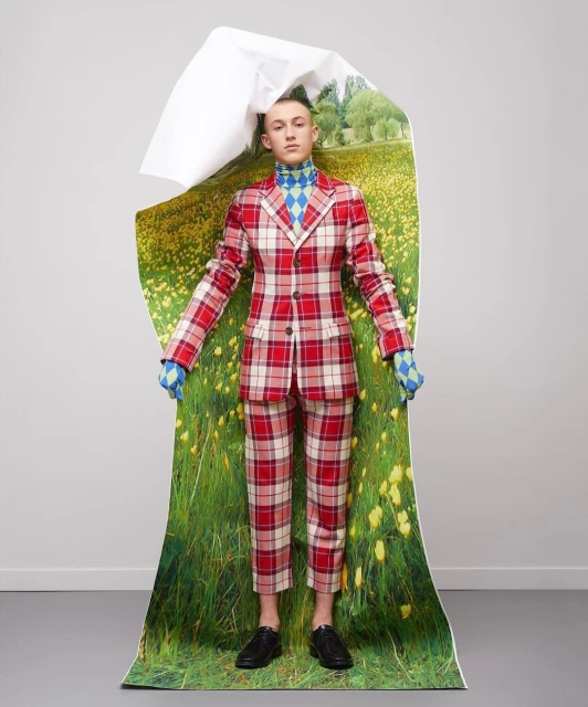 A person stands indoors wearing a red plaid suit, a blue argyle turtleneck, and black shoes. Behind them is a torn field image.