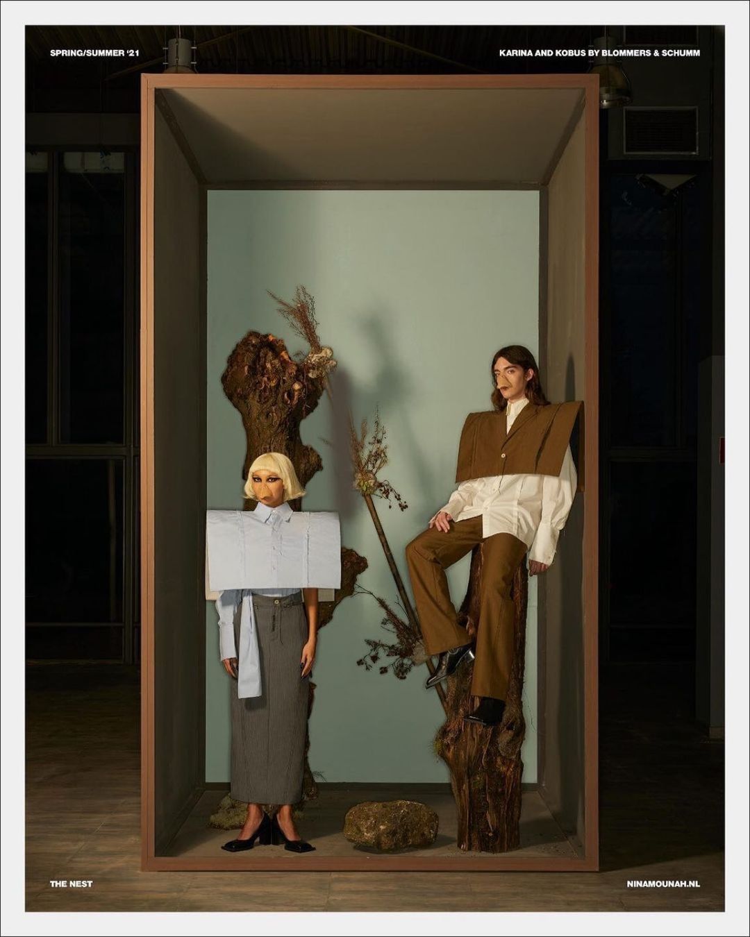 Two women pose in a constructed box-like display in a studio. One sits on a wooden stump, the other stands in a gray skirt and large square top.