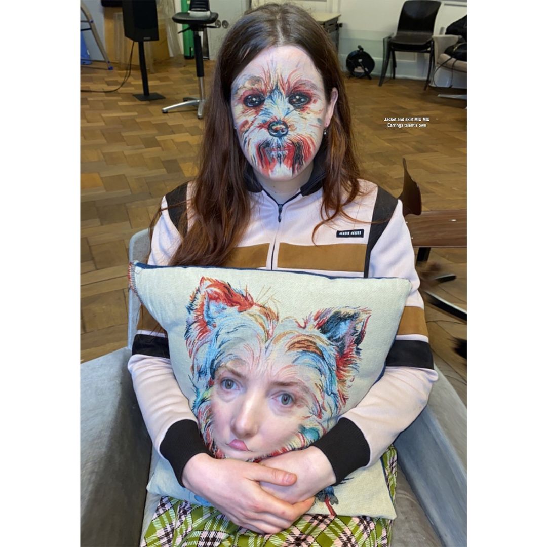 A woman with dog face paint sits, holding a pillow with a woman/dog face mashup, indoors with chairs and wood floor visible.