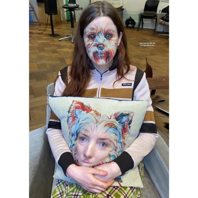 A woman with dog face paint sits, holding a pillow with a woman/dog face mashup, indoors with chairs and wood floor visible.