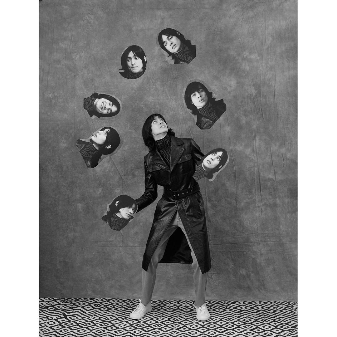 A person in a black coat surrounded by floating headshot photos against a textured backdrop and geometric patterned floor.