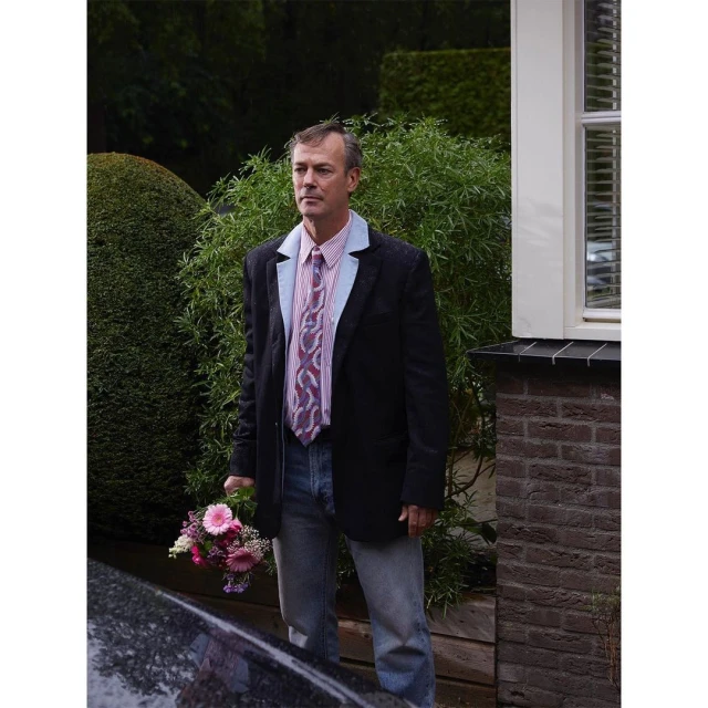 Mature man in a blazer, light jeans, and a tie holds a colorful bouquet outdoors near a brick wall, with green plants in the background.