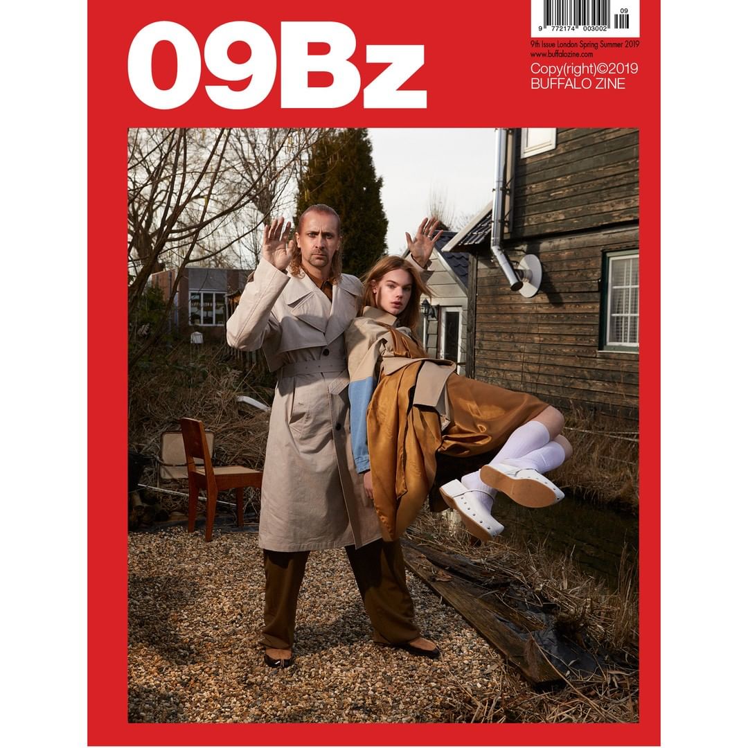 Fashion magazine cover featuring a man and woman in trench coats standing outdoors in a rural setting with a red border.