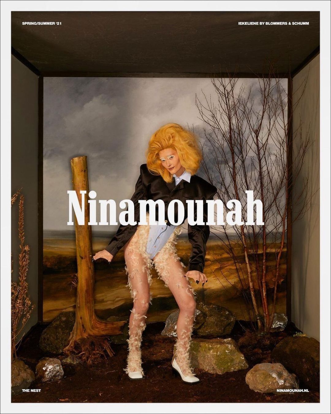 Fashion editorial shot of a stylish woman in a faux fur outfit posing in a box-like studio setting with a painted backdrop and props.