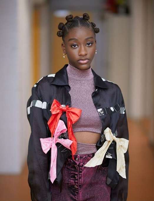 Portrait of a woman with space buns wearing a black jacket with pink, red, and white bows posing indoors with a neutral expression.