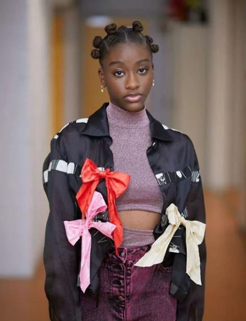 Portrait of a woman with space buns wearing a black jacket with pink, red, and white bows posing indoors with a neutral expression.