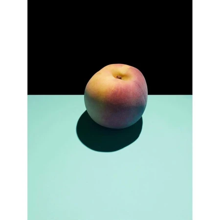 A ripe peach sits on a flat turquoise surface against a stark black background, creating contrast and highlighting its colors and form.