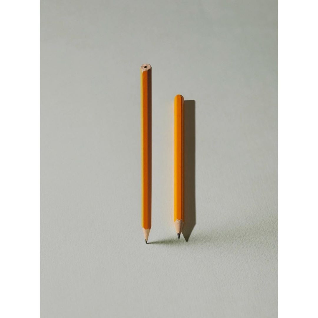 Two yellow pencils stand on a light gray surface, one sharpened and the other unsharpened, casting soft shadows in a simple composition.
