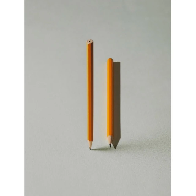 Two yellow pencils stand on a light gray surface, one sharpened and the other unsharpened, casting soft shadows in a simple composition.