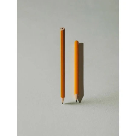 Two yellow pencils stand on a light gray surface, one sharpened and the other unsharpened, casting soft shadows in a simple composition.