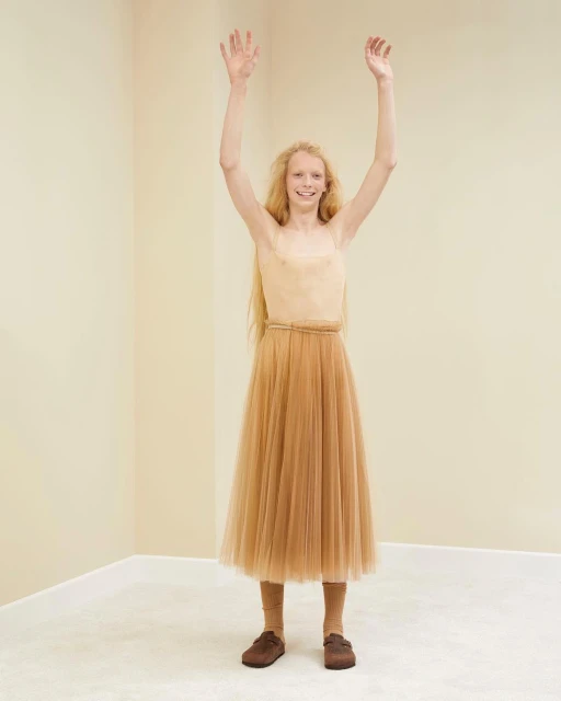 Young woman in a beige top and tulle skirt stands with her arms raised in an indoor corner, showcasing a fashion-forward and joyful pose.