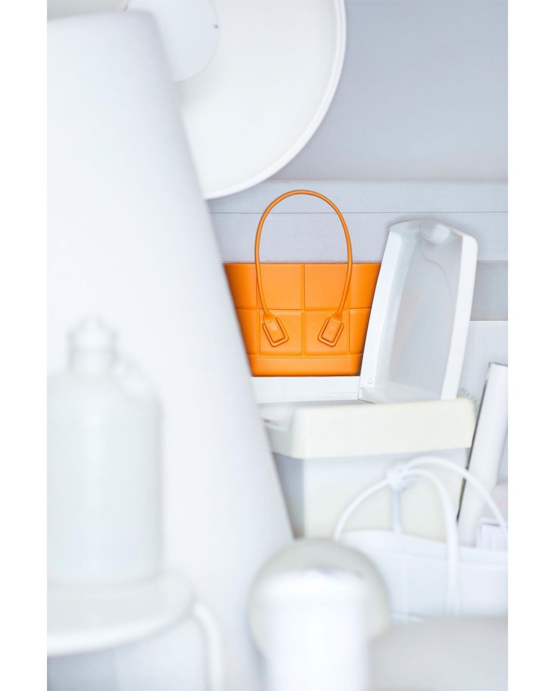 An orange leather handbag with top handles is vertically oriented in an eye-level shot, surrounded by white props in a neutral setting.